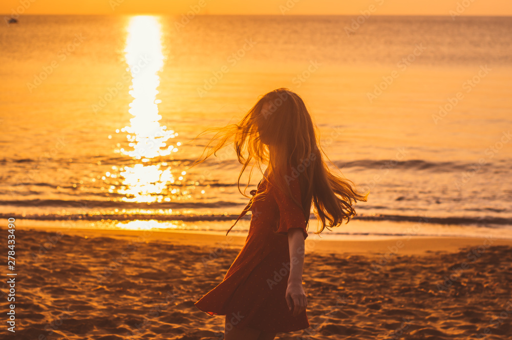Young blonde girl in red dress dancing in the evening on the beach