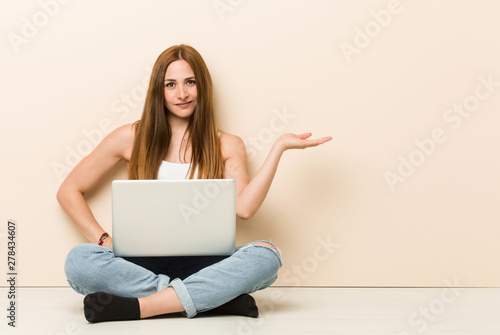 Young ginger woman sitting on her house floor showing a copy space on a palm and holding another hand on waist. © Asier
