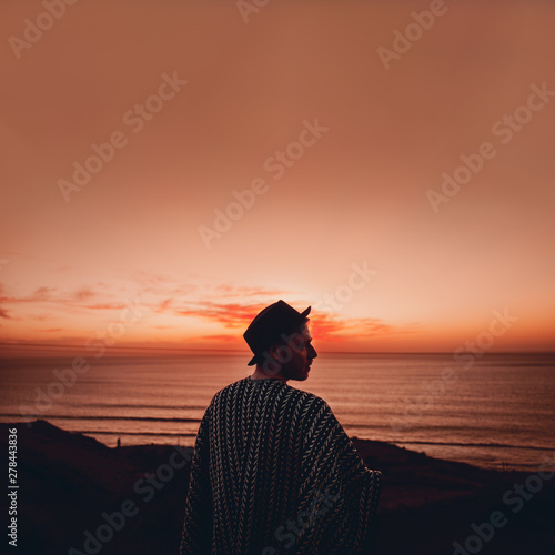 Mann mit Poncho und Hut blickt bei Sonnenuntergang auf das Meer in Marokko photo