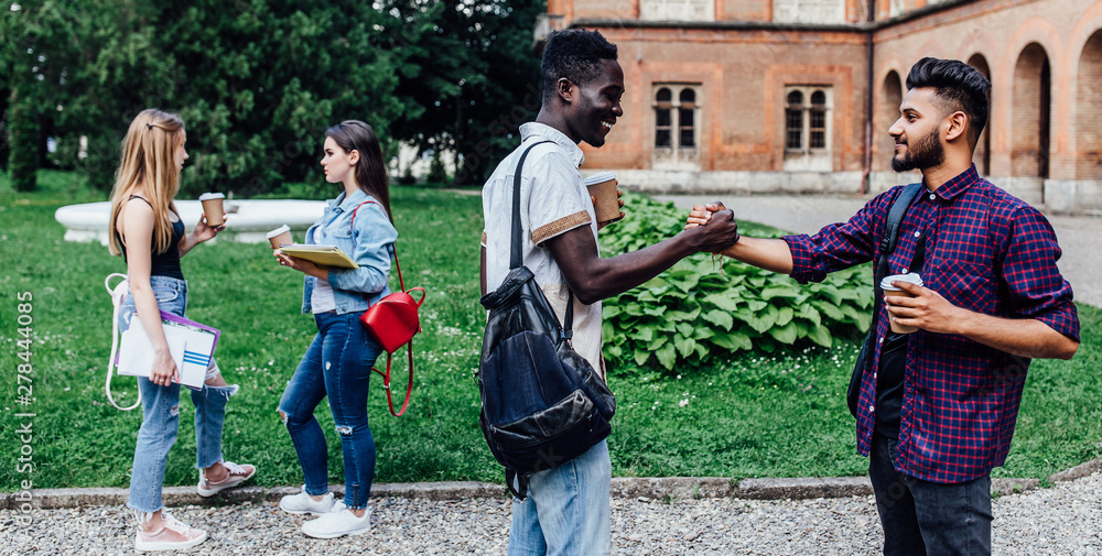 Fototapeta premium Meet of students. Two handsome young male student holding coffe, have break time while walking on green grass near university.