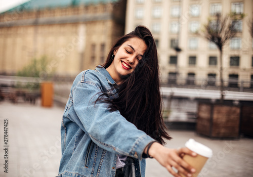 Outdoor portrait of a dark-haired young woman in a denim jacket holds coffee to go. Emotional woman posing with happy smile on the street.