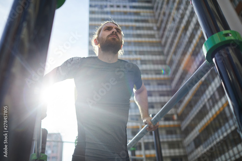 Below view of determined bearded athlete in tshirt hanging on parallel bars and looking into distance on outdoor ground
