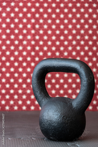 Fototapeta Naklejka Na Ścianę i Meble -  Rustic black kettlebell on a black rubber mat floor against a patterned backdrop of red and white