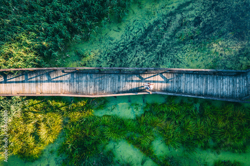 Aerial top view of woman traveller on wooden bridge pathway over marshy river with vegetation thickets, summer travel concept