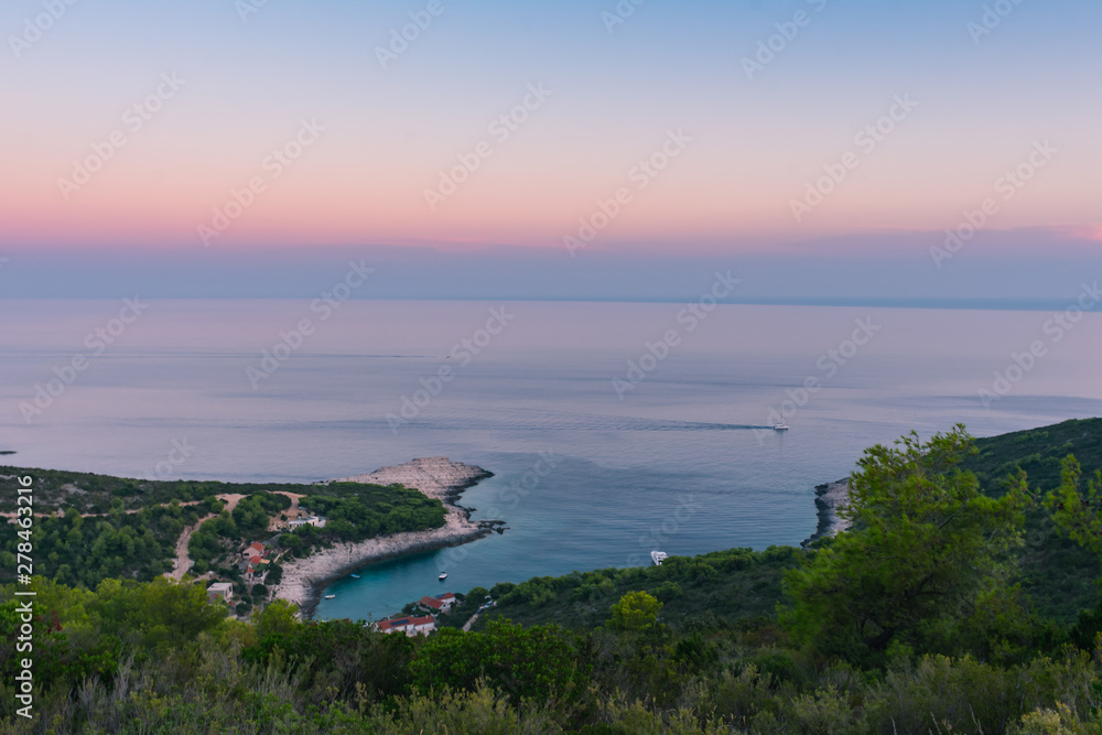 Landscape view of a beautiful blue bay in the Adriatic sea, Vis island in Croatia, viewed from above over green mediterranean vegetation, with moored sailing boats and houses during a colorful sunset 