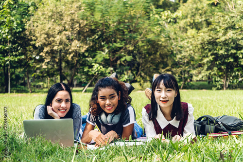 Group of smiling international students or teenagers using laptop computer doing their homework with book together in park at university.Education and friendship Concept