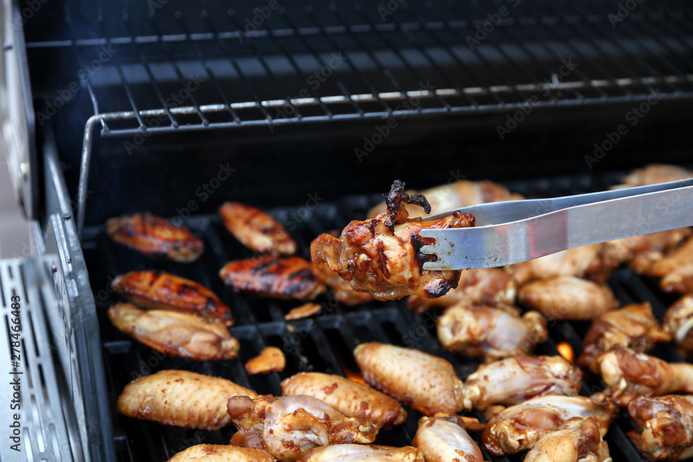 Close up of an open barbecue grill cooking chicken wings with tong holding a single wing Close up of an open barbecue grill cooking chicken wings with tong holding a single wing