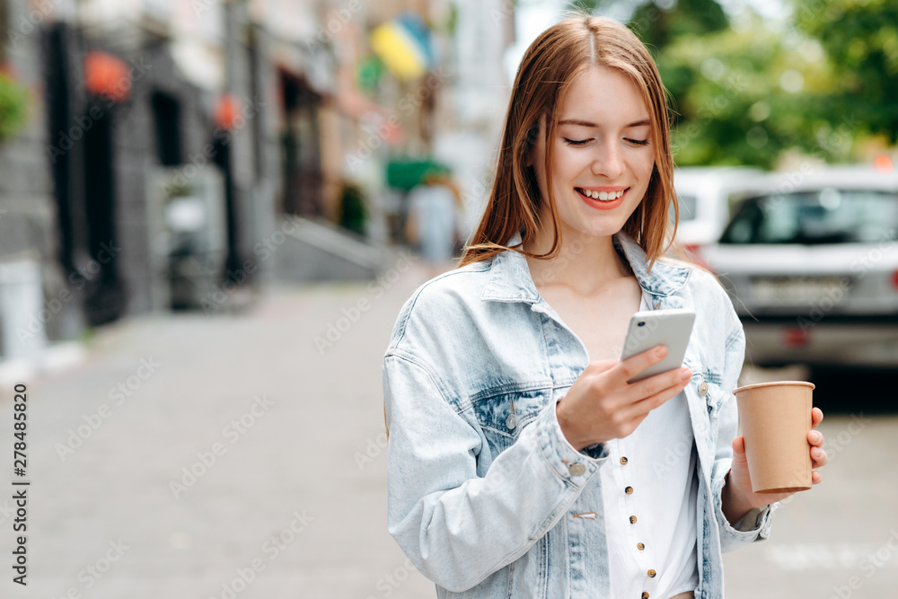 Happy young woman standing and holding a smartphone looking at the screen outdoor- Image Happy young woman standing and holding a smartphone looking at the screen outdoor- Image