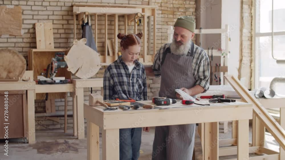 Following shot of Caucasian aged hipster man and his granddaughter coming to workbench in workshop, man putting on glasses and starting talking about his craft and showing tools to girl Following shot of Caucasian aged hipster man and his granddaughter coming to workbench in workshop, man putting on glasses and starting talking about his craft and showing tools to girl