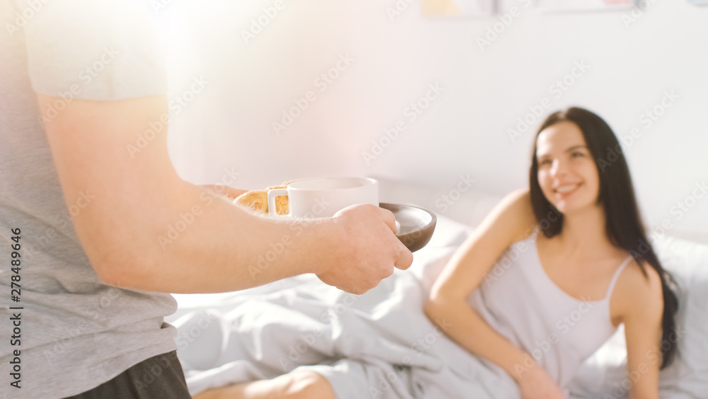 Shot of a Caring Young Man Bringing Tray with Breakfast in Bed to His Lovely Female Partner. Ideal Tender Relationship and Coffee and Croissants in Bed. Shot with Warm Sunlight. Shot of a Caring Young Man Bringing Tray with Breakfast in Bed to His Lovely Female Partner. Ideal Tender Relationship and Coffee and Croissants in Bed. Shot with Warm Sunlight.
