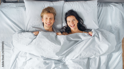 Happy and Fun Young Couple Lying under Blanket on a Bed, Making Silly Faces and Laughing. Lovely Young People having Fun. Top Down Shot.