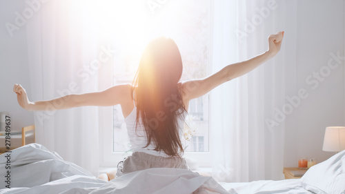 Beautiful Brunette is Waking up in the Morning, Stretches in the Bed, Sun Shines on Her From the Big Window. Happy Young Girl Greets New Day with Warm Sunlight Flare.