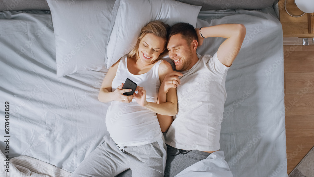 Obraz premium Loving Young Couple Spending Morning in Bed, Pregnant Young Woman Shows Her Partner Something on a Touchscreen Smartphone. Searching for Baby Clothes, Browsing Through Social Media, Posting Pictures.