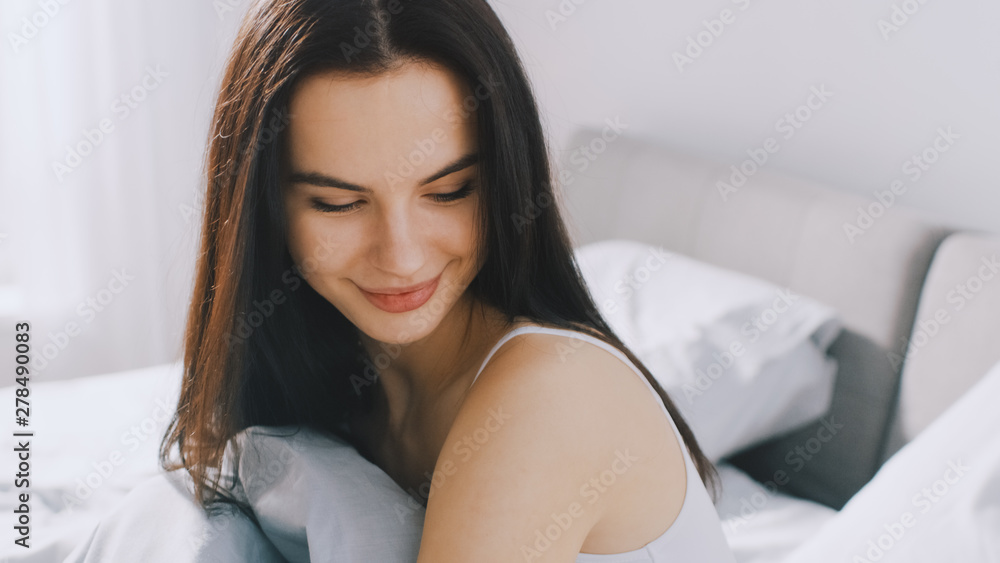 Beautiful Brunette Slowly Waking up in the Morning, Sits Up on the Bed, Smiles Charmingly. Sun Shines on a Happy Young Girl From the Big Window. Beautiful Brunette Slowly Waking up in the Morning, Sits Up on the Bed, Smiles Charmingly. Sun Shines on a Happy Young Girl From the Big Window.
