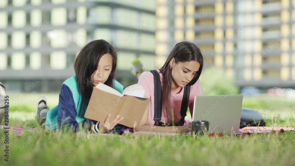 Two diverse female college students lying on blanket on grass and doing project together, Caucasian girl typing on laptop, her Asian friend reading textbook Two diverse female college students lying on blanket on grass and doing project together, Caucasian girl typing on laptop, her Asian friend reading textbook