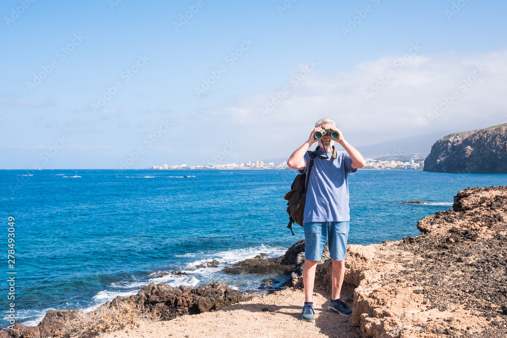 Senior people with white hair standing on cliff at the ocean. Casual clothing with backpack and looking with binoculars. Sunlight and seascape