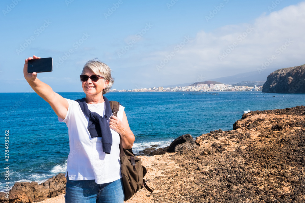 Smiling senior woman standing on the cliff over the sea. Blue sky and water. Casual clothing with backpack. Takes a selfie with phone. Freedom and relaxation