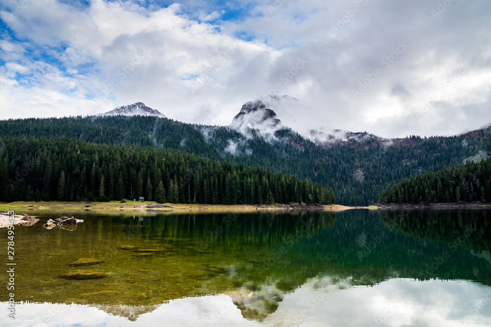 Montenegro, Reflecting calm waters of black lake in durmitor national park surrounded by green forest and high snow covered mountains partly hidden in fog near zabljak