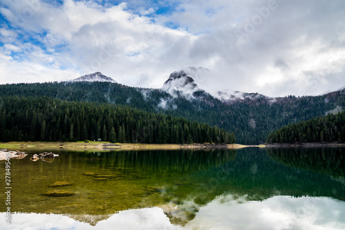 Montenegro, Reflecting calm waters of black lake in durmitor national park surrounded by green forest and high snow covered mountains partly hidden in fog near zabljak © Simon