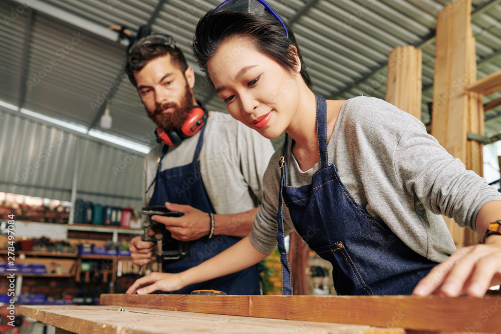Smiling Vietnamese female carpenter working with wood under control of coworker Smiling Vietnamese female carpenter working with wood under control of coworker