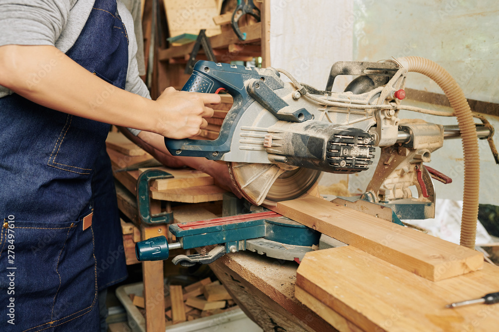 Cropped image of carpenter in denim overall cutting wooden board when working alone in studio Cropped image of carpenter in denim overall cutting wooden board when working alone in studio