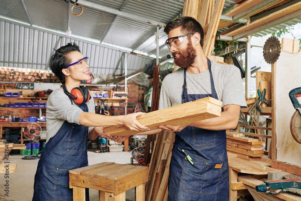 Smiling female carpenter in denim apron giving stack of wooden planks to her coworker Smiling female carpenter in denim apron giving stack of wooden planks to her coworker