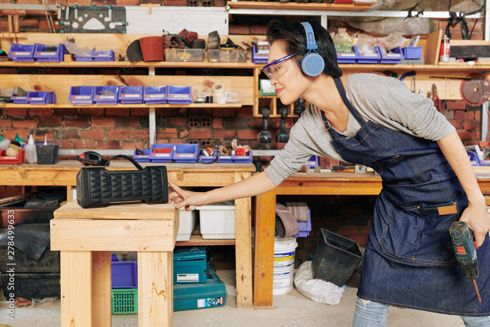 Smiling female carpenter tuning on speaker before starting to work with wood Smiling female carpenter tuning on speaker before starting to work with wood
