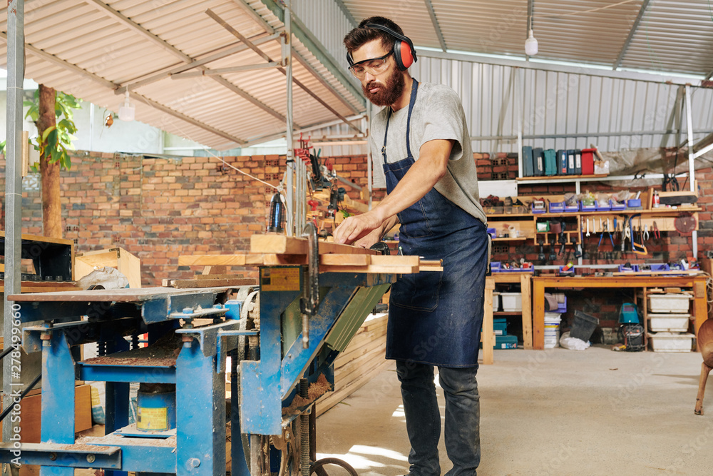 Serious Caucasian carpenter wearing ear muffs when cutting wooden plank on workbench with circular saw
