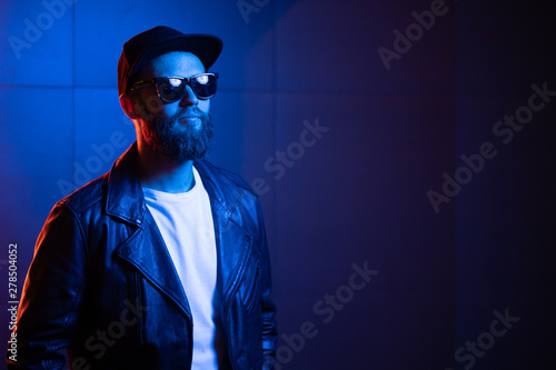 Hipster handsome man on the city streets being illuminated by neon signs. He is wearing leather biker jacket or asymmetric zip jacket with black cap, jeans and sunglasses.