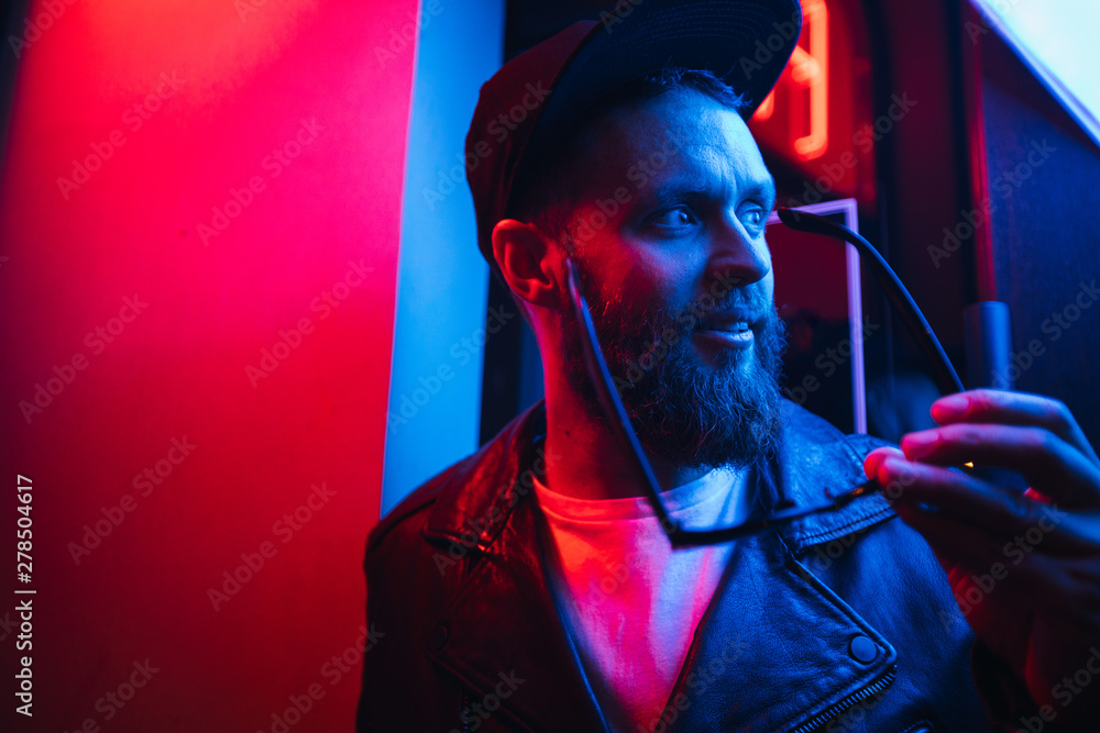 Hipster handsome man on the city streets being illuminated by neon signs. He is wearing leather biker jacket or asymmetric zip jacket with black cap, jeans and sunglasses. - obrazy, fototapety, plakaty 