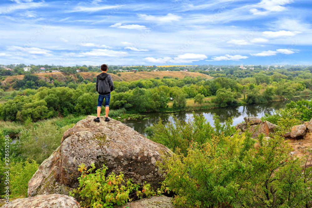 A teenager stands on top of a large stone boulder on the banks of the Southern Bug and looks into the distance.