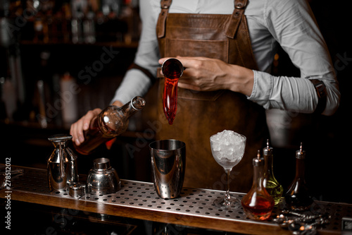 Male bartender pouring a red alcoholic drink from the steel jigger to the cocktail shaker