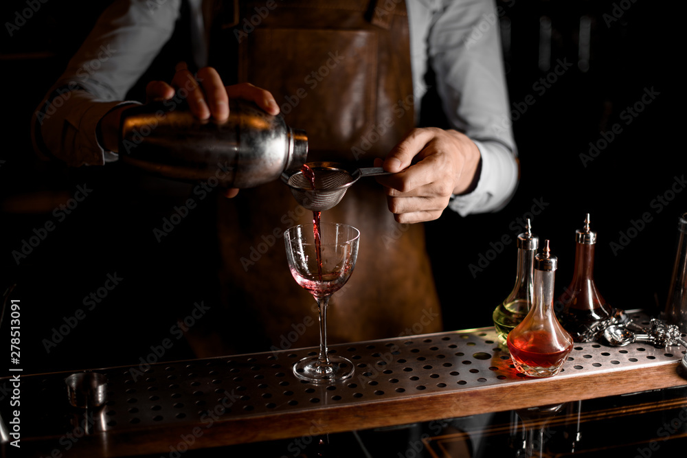 Male bartender pouring a red alcoholic drink from the steel shaker through the sieve