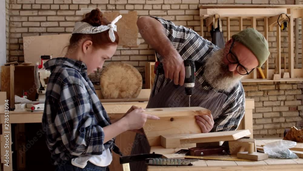 Medium shot of Caucasian ginger girl holding wooden planks while aged hipster man screwing screw in them using electrical screwdriver in carpentry shop Medium shot of Caucasian ginger girl holding wooden planks while aged hipster man screwing screw in them using electrical screwdriver in carpentry shop