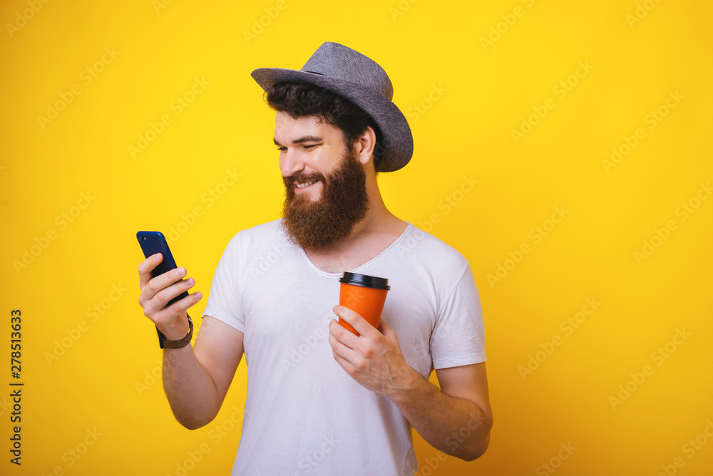 Portrait of handsoema bearded guy in hat, using his mobile phone while holding a cup with coffee, standing over yellow background Portrait of handsoema bearded guy in hat, using his mobile phone while holding a cup with coffee, standing over yellow background
