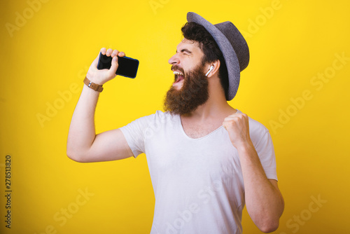 Handsomea excited bearded guy, wearing a hat,  holding a mobile phone, and enjoying music listening on earphones, over isolated yellow background