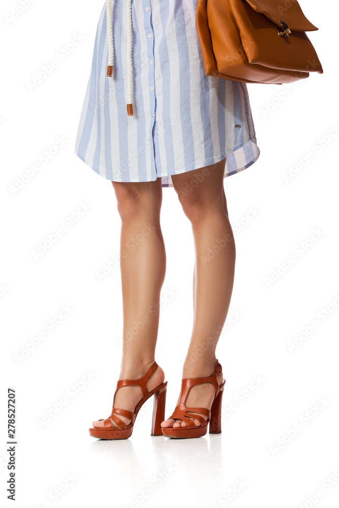 Brown women's shoes and leather bag. Beautiful female legs wearing summer high heeled sandals in blue dress and brown woman handbag on white background.