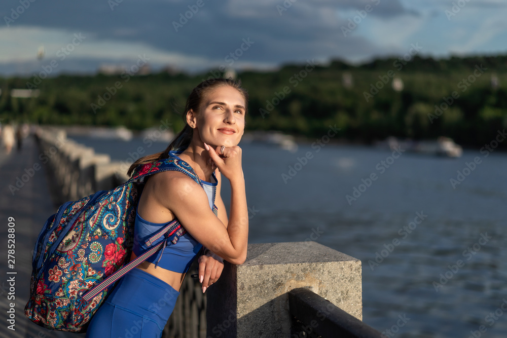 girl walks at sunset along the embankment in a blue sports suit with a colored backpack. Summer evening young athlete resting on the embankment of the river.