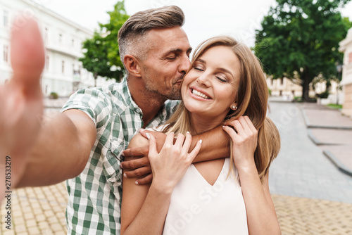 Image of loving young couple kissing and taking selfie photo together while walking through city street