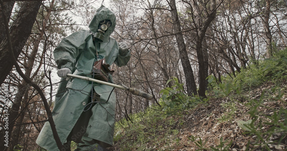 Low angle unrecognizable guy in hazmat suit and gas mask searching for radiation with Geiger counter in abandoned forest near Chernobyl.