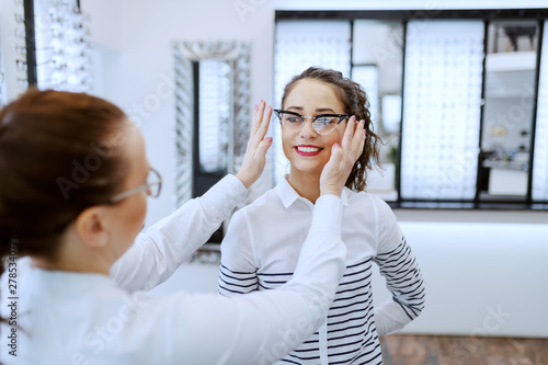 Female optician putting eyeglasses on beautiful Caucasian smiling brunette. Optician shop interior.