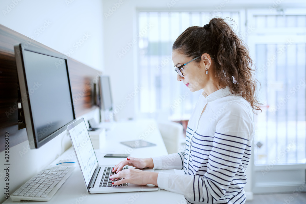 Side view of hard working Caucasian businesswoman with brown curly hair and eyeglasses typing on laptop. Office interior. Side view of hard working Caucasian businesswoman with brown curly hair and eyeglasses typing on laptop. Office interior.