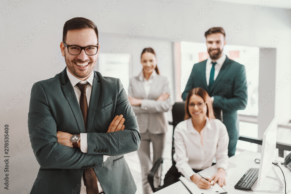 Young handsome smiling CEO in suit standing in office with arms crossed. In background his successful team posing. Young handsome smiling CEO in suit standing in office with arms crossed. In background his successful team posing.