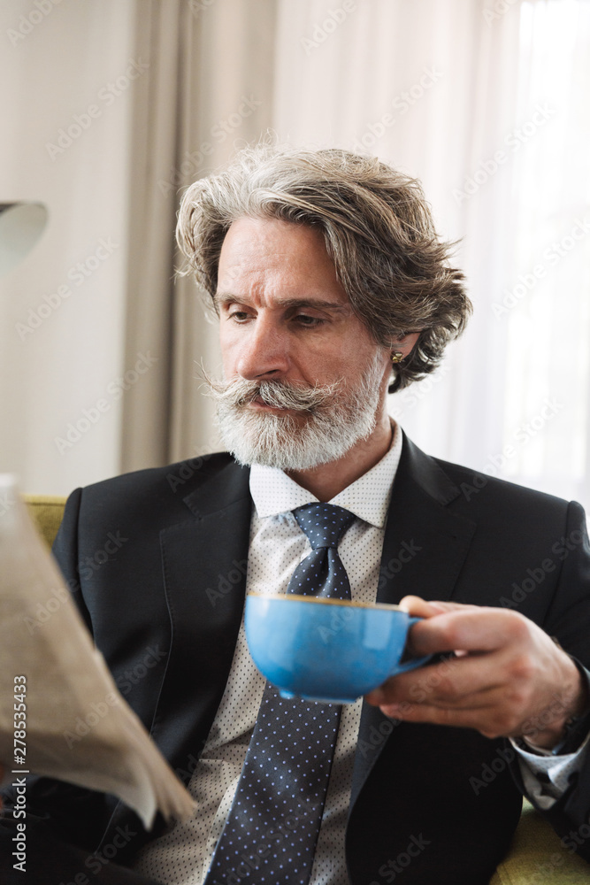 Concentrated senior gray-haired business man sitting in chair indoors at home reading newspaper drinking coffee. Concentrated senior gray-haired business man sitting in chair indoors at home reading newspaper drinking coffee.