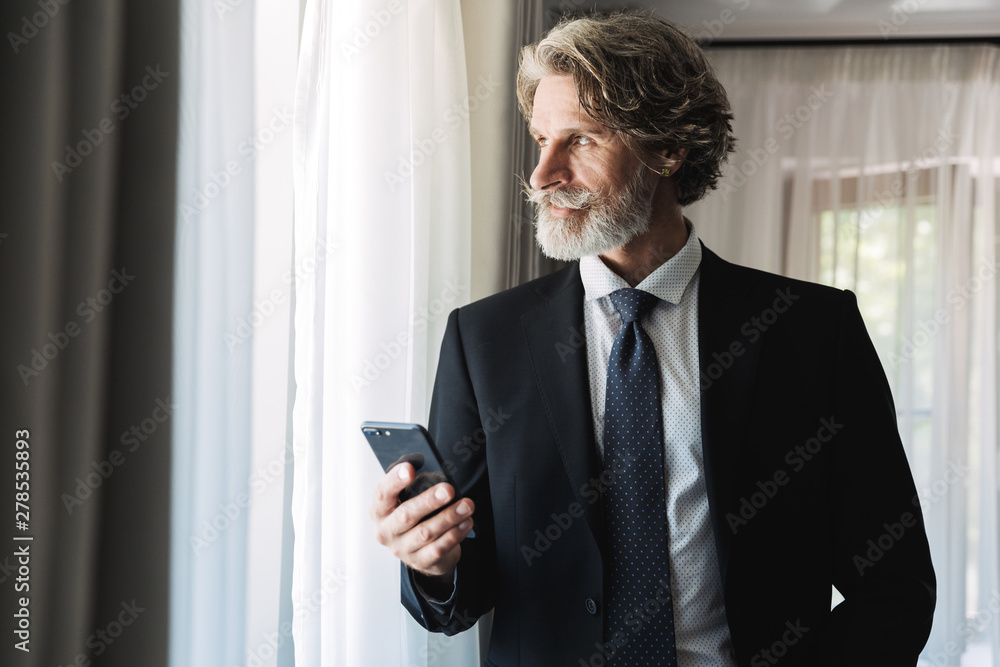 Image of unshaven adult businessman wearing black suit holding smartphone in hotel apartment Image of unshaven adult businessman wearing black suit holding smartphone in hotel apartment
