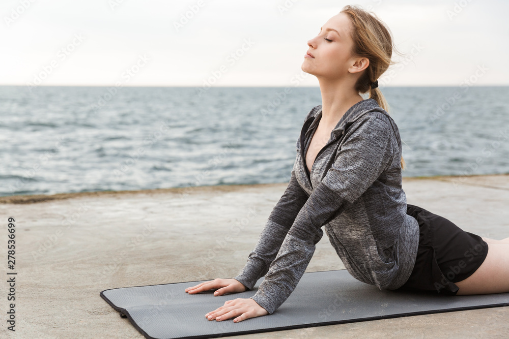 Image of cute concentrated woman doing exercise on mat while working out on pier near seaside in morning