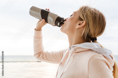 Image of young woman drinking water from bottle while working out on pier near seaside in morning