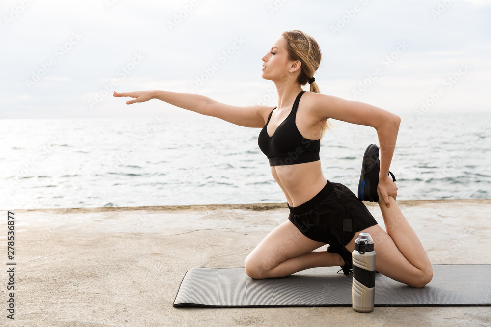 Image of seductive woman doing exercise on mat with water bottle while working out near seaside in morning Image of seductive woman doing exercise on mat with water bottle while working out near seaside in morning