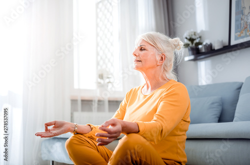 Slika na platnu Senior female in lotus pose meditating sitting on floor at home