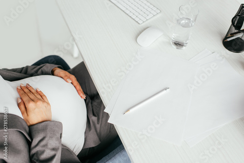 cropped view of pregnant woman sitting behind table with glass, keyboard, papers and pencil